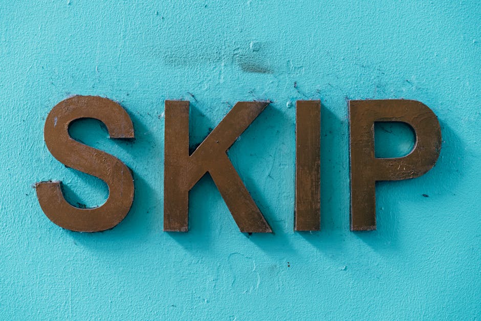 Close-up view of three large, three-dimensional metal letters spelling 'SKIP' mounted on a smooth, light blue painted wall. The letters have a matte brown finish, with visible surface textures showing