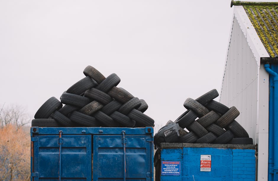 The image depicts a collection of overflowing rubbish bins and discarded waste materials situated on a paved area adjacent to a car parking space. The central focus is on a large grey mixed paper and