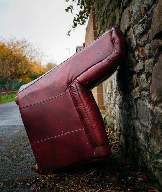A red leather armchair with visible creases and a smooth, slightly glossy surface is shown discarded on a damp outdoor pavement, leaning against a rough, dark stone wall composed of irregularly shaped