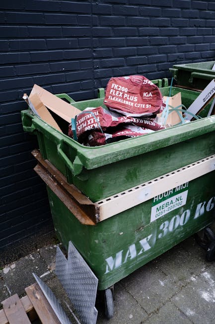 A red leather armchair with visible creases and a smooth, slightly glossy surface is shown discarded on a damp outdoor pavement, leaning against a rough, dark stone wall composed of irregularly shaped