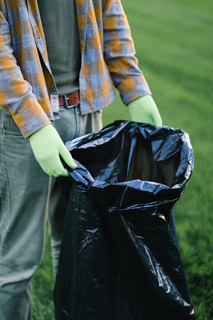 A worker wearing a blue cap, an orange high-visibility vest, and blue uniform bins out rubbish into a blue wheelie bin beside a white waste collection truck parked on a narrow cobblestone street in an